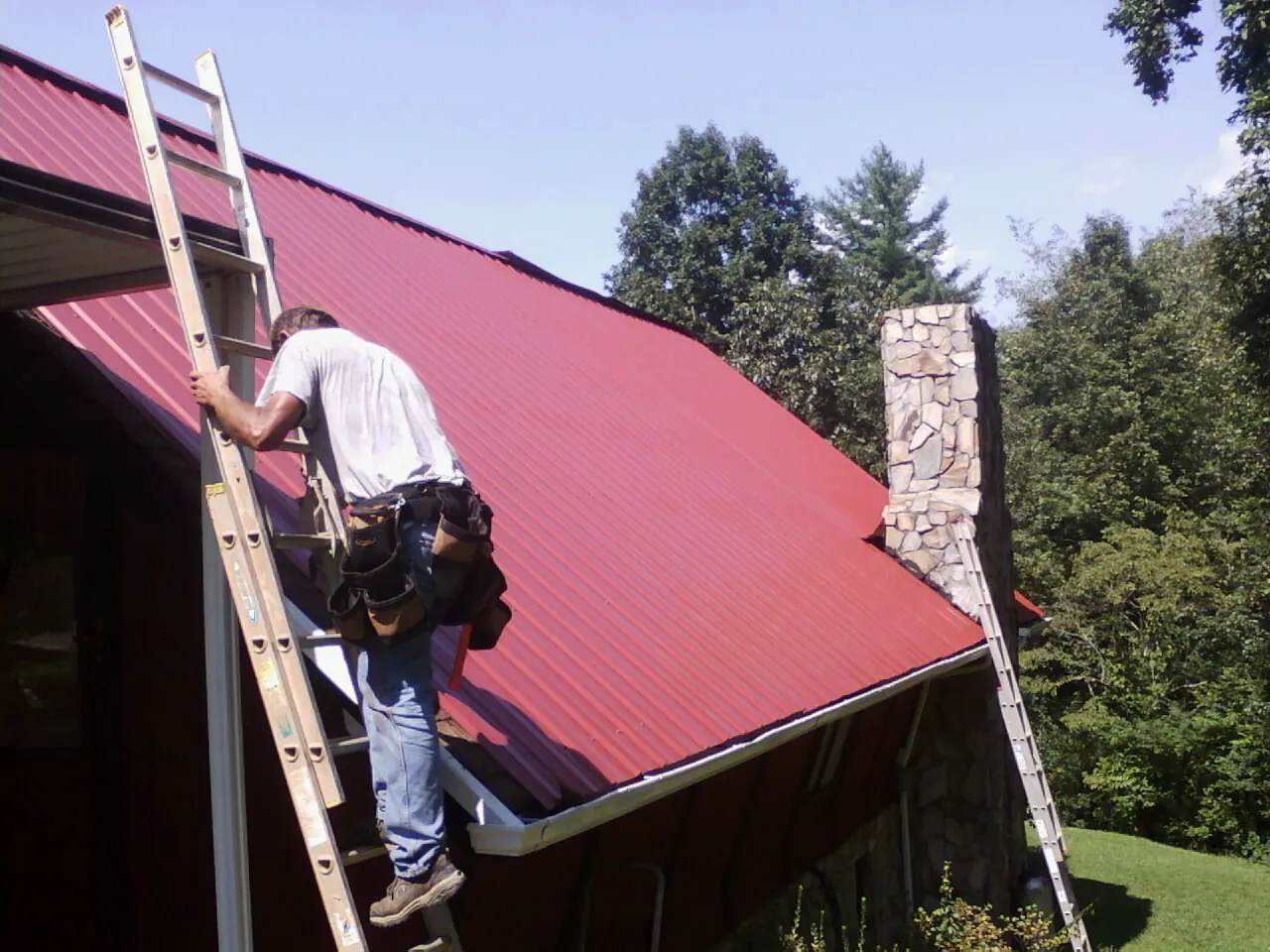 Roofer on a ladder beside a red metal roof and stone chimney — Soffit & Fascia Repair by Roofing Brothers in Oshkosh